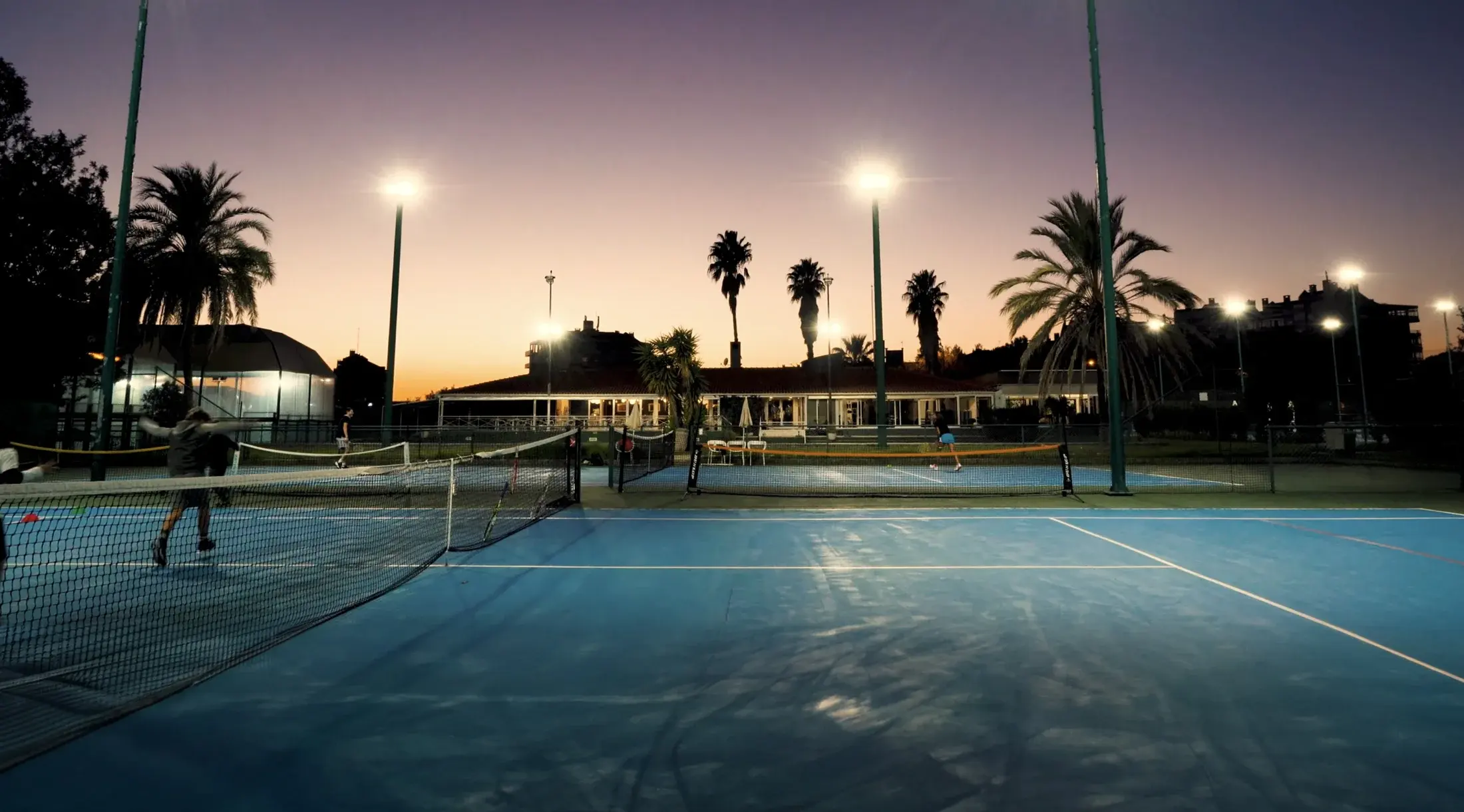 Quadras de tênis externas de piso duro iluminadas, sede do clube e palmeiras no Lisboa Racket Centre ao anoitecer